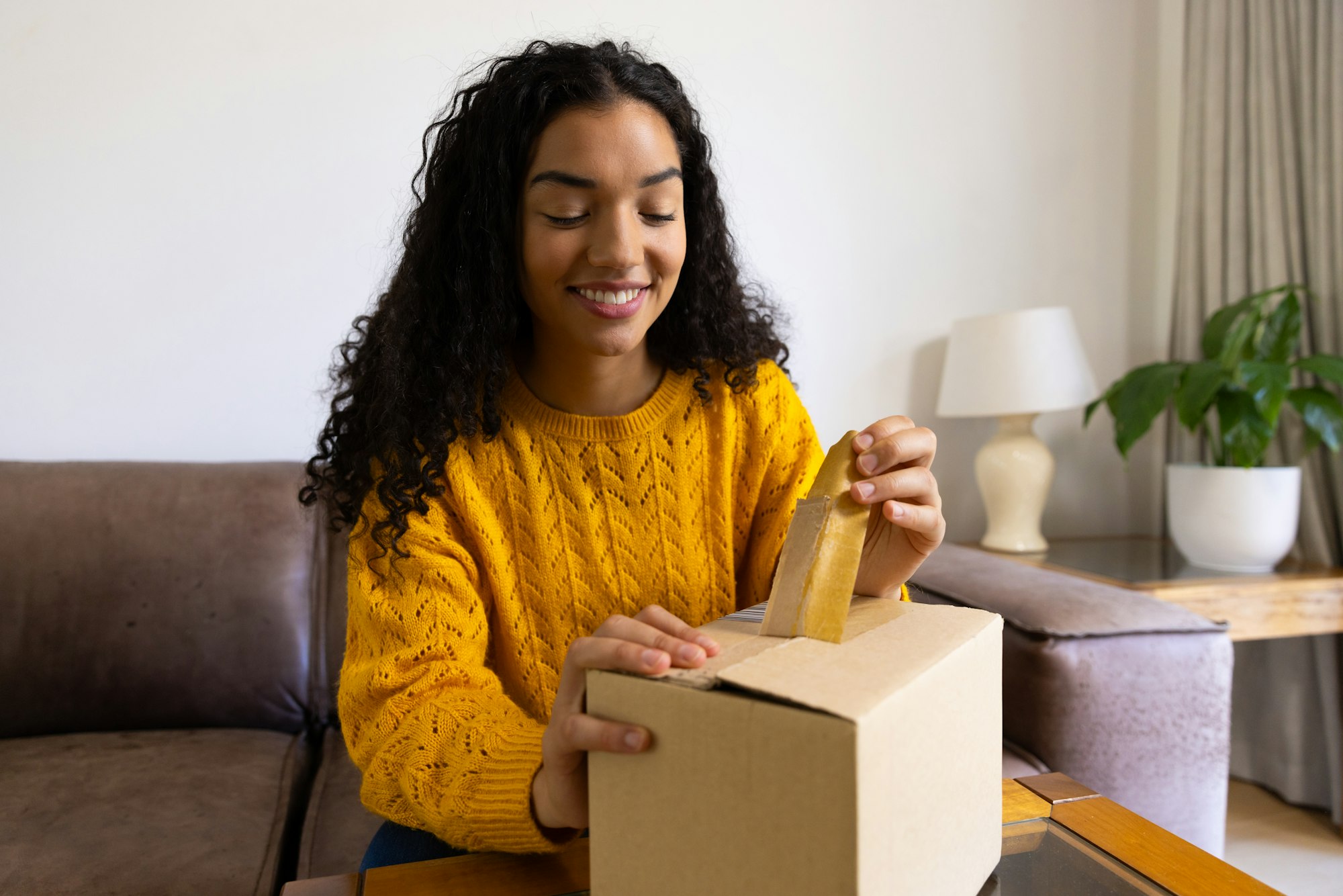 Happy biracial woman in yellow sweater opening package at home